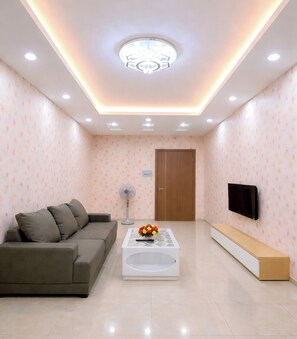 A modern living room with a sofa, coffee table, TV, and recessed ceiling lighting. - HQH Sea View Apartment (Bac Nha Trang)