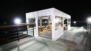 Una estructura blanca semblant a una tenda amb una taula i caires a l'interior, situada en una terrassa amb baranes metàl·liques.