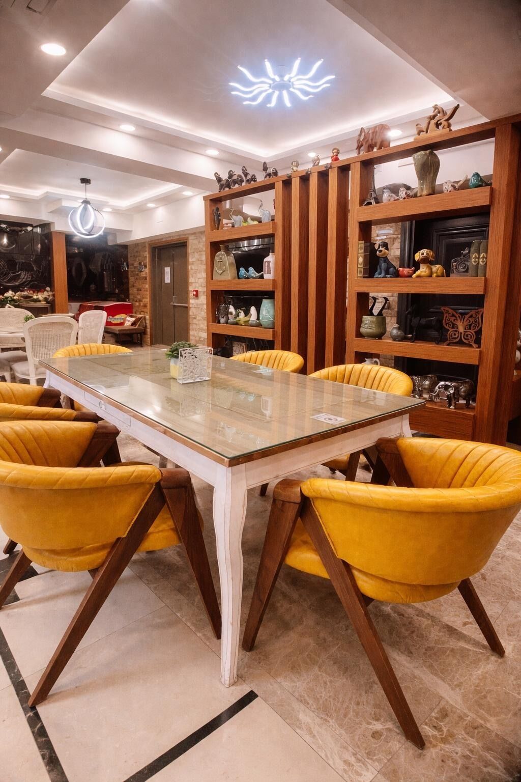 Un comedor con mesa de vidrio, sillas tapizadas de color amarillo, estanterías de madera con adornos y una lámpara en el techo.