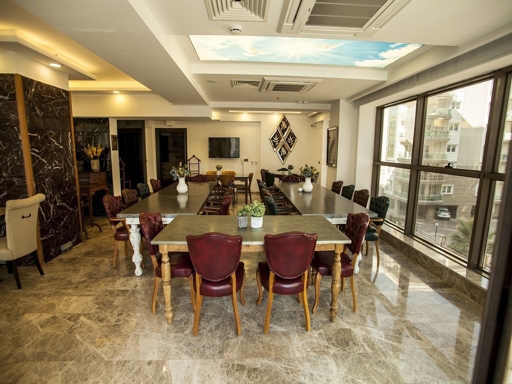 Un comedor amplio con una mesa larga, sillas tapizadas en rojo y vistas a edificios a través de grandes ventanales.