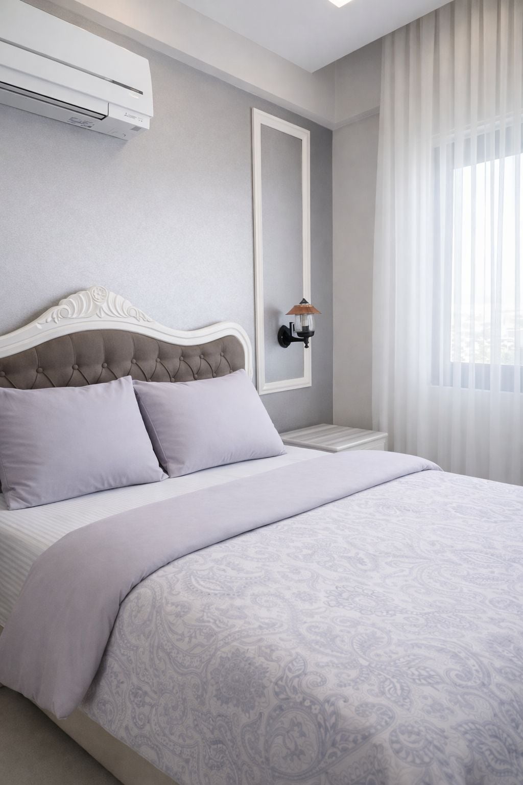 Un dormitorio con cabecera capitonada, cama con ropa de cama estampada, mesita de noche, lámpara de pared y unidad de aire acondicionado.