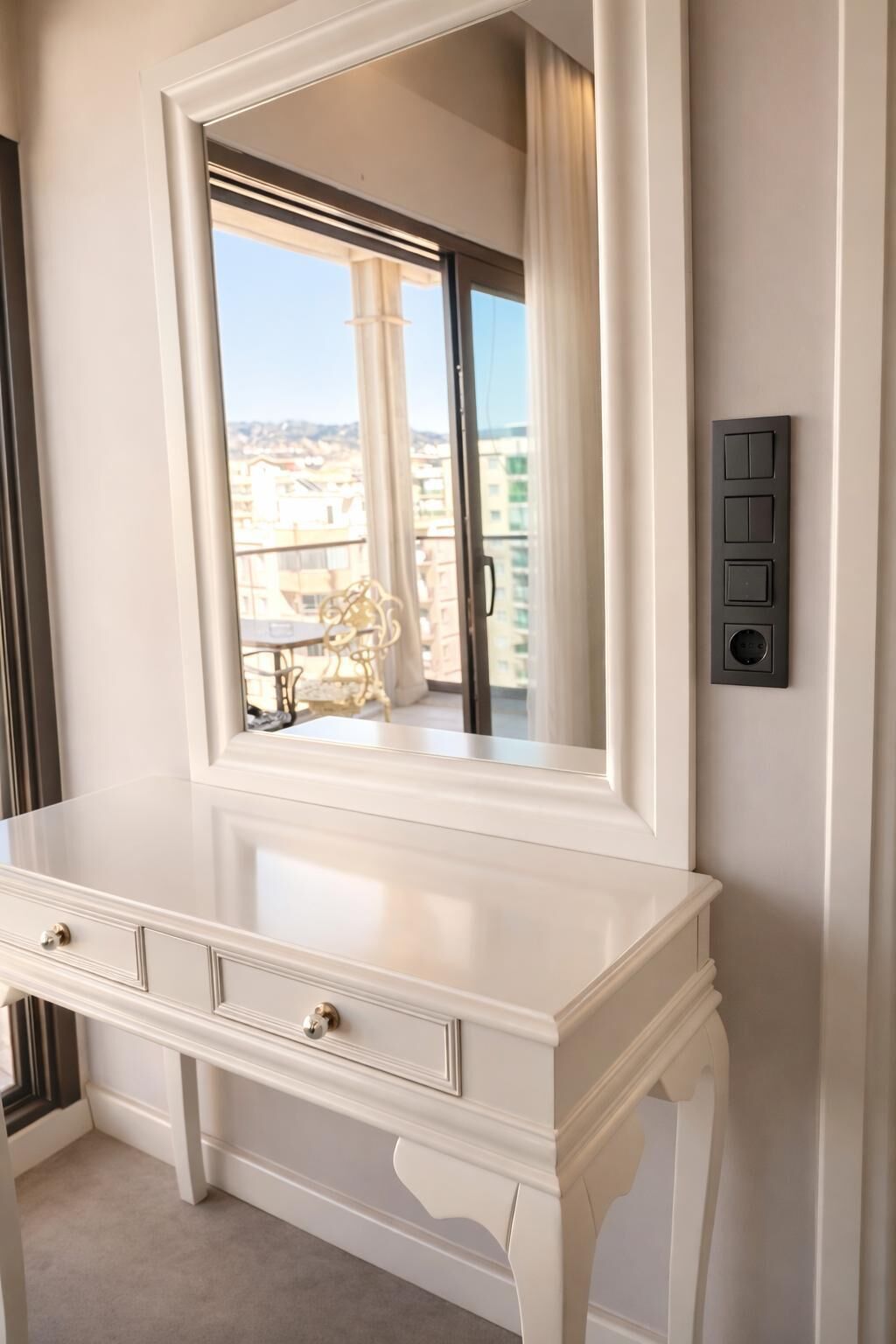 A white vanity with a large mirror, a wall-mounted light switch, and a view of buildings through the window.