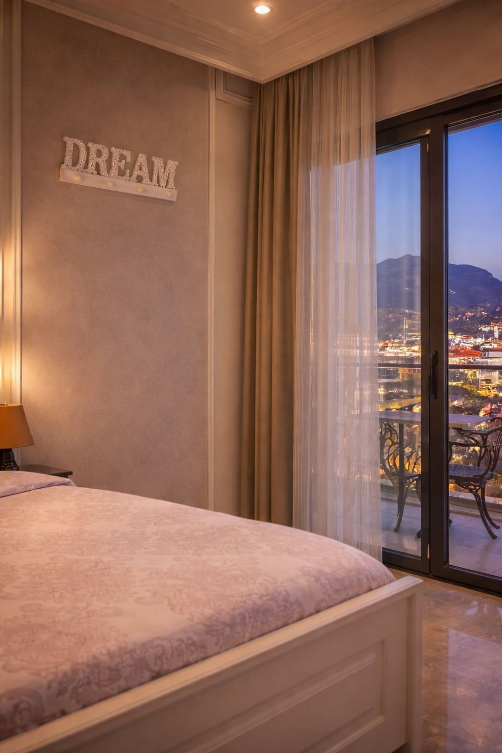 Een slaapkamer met een bed, een 'DREAM' bord, uitzicht op een stadsgezicht door een glazen deur en een balkon met stoelen.