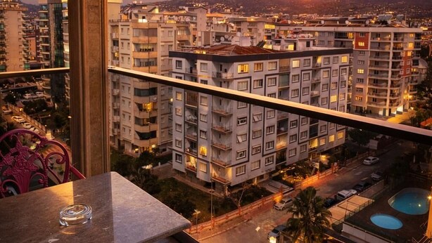 Un balcón con vistas a un paisaje urbano al atardecer, una piscina y un área de comedor con sillas.