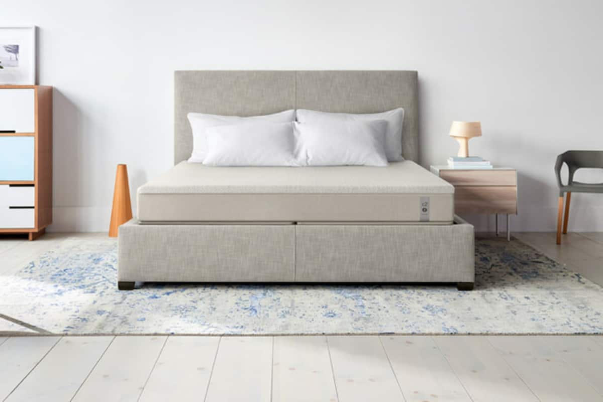 A bed with a grey headboard and two pillows, a bedside table with a lamp, a chair, and a small wooden cabinet.