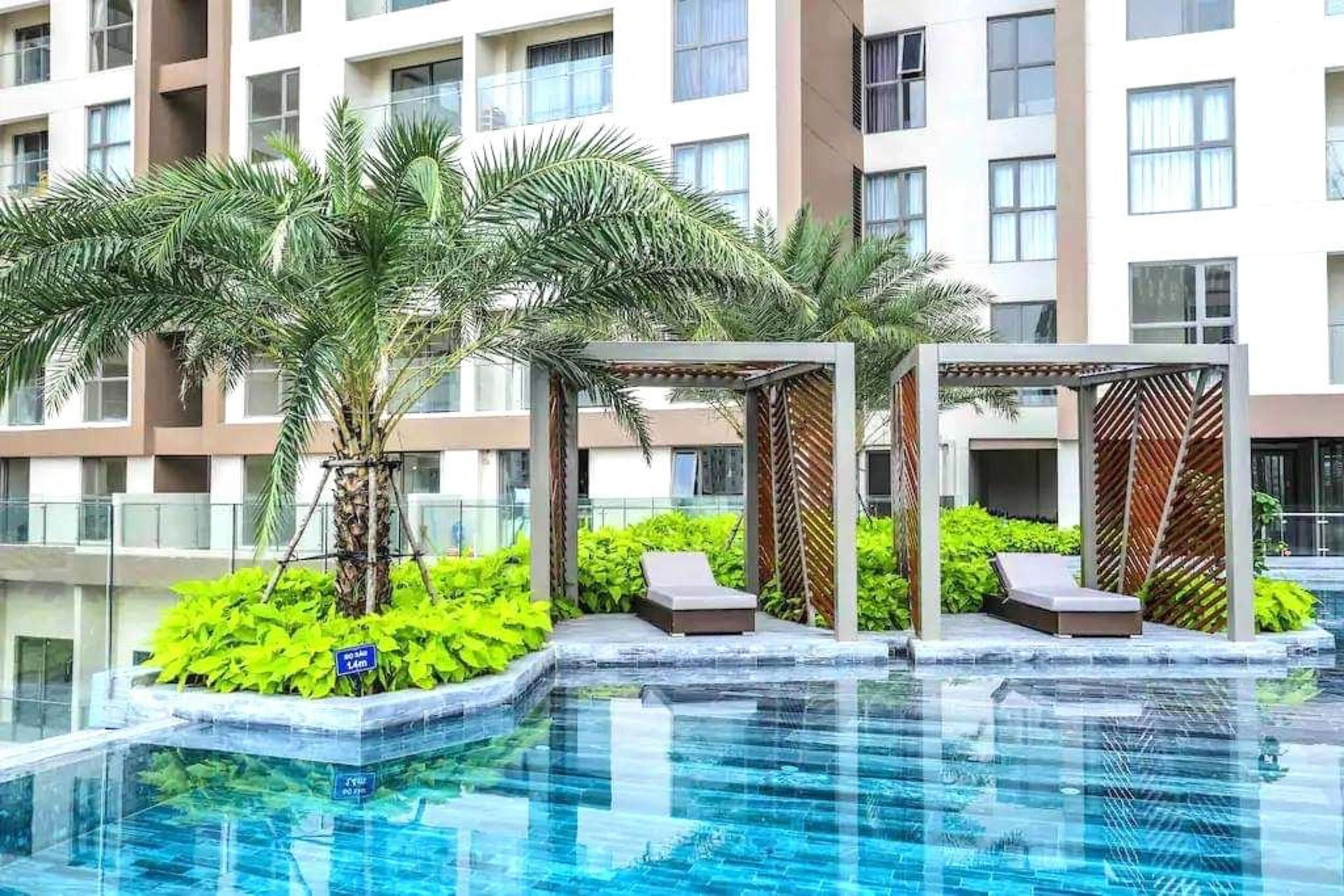 A swimming pool with palm trees and lounge chairs, surrounded by a modern building with multiple windows.