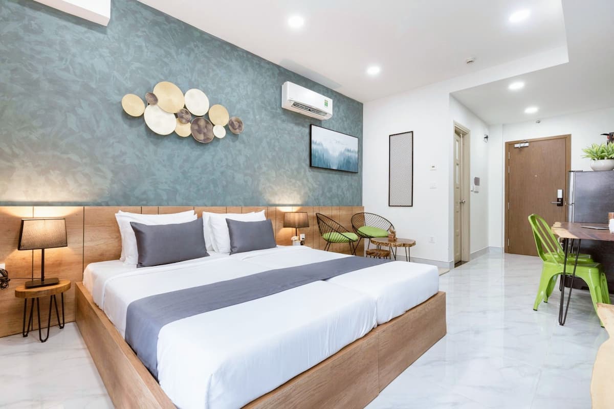 A modern hotel room with a large bed, a wooden headboard, a wall-mounted air conditioner, a framed picture, and a dining area with a table and chairs.