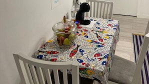 Ein Essbereich mit einem Tisch, der mit einer bunten Tischdecke bedeckt ist, weißen Stühlen, einer Obstschale und einer schwarzen Katze, die auf dem Tisch sitzt.