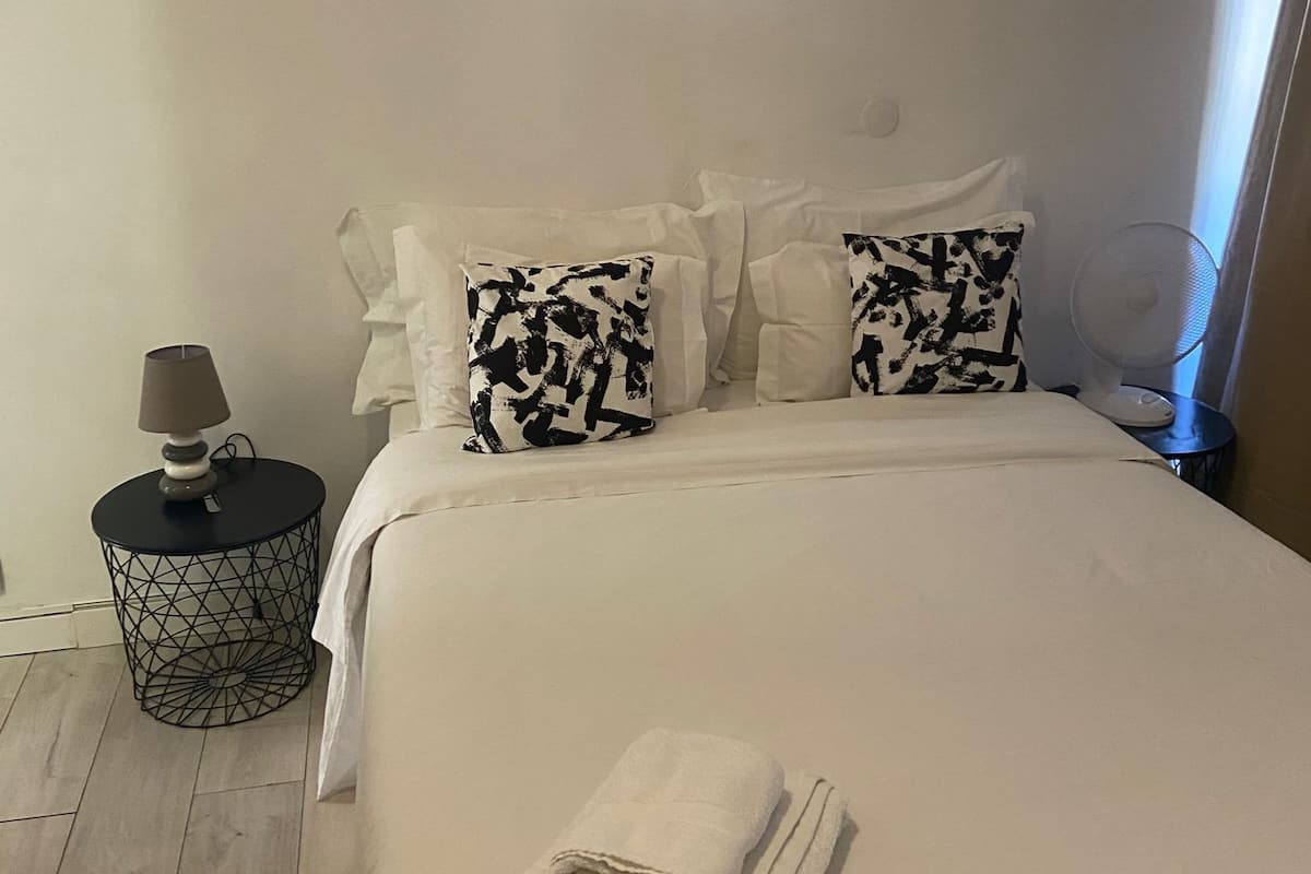 Een net opgemaakt bed met witte beddengoed, twee gestreepte kussens, een klein rond nachtkastje met een lamp, en een opgevouwen handdoek op het bed.