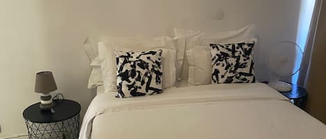 A neatly made bed with white bedding, two patterned pillows, a small round side table with a lamp, and a folded towel on the bed.