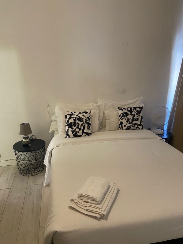 A neatly made bed with white bedding, two patterned pillows, a small round side table with a lamp, and a folded towel on the bed. - Santo Amaro Guest House (Lisbon)