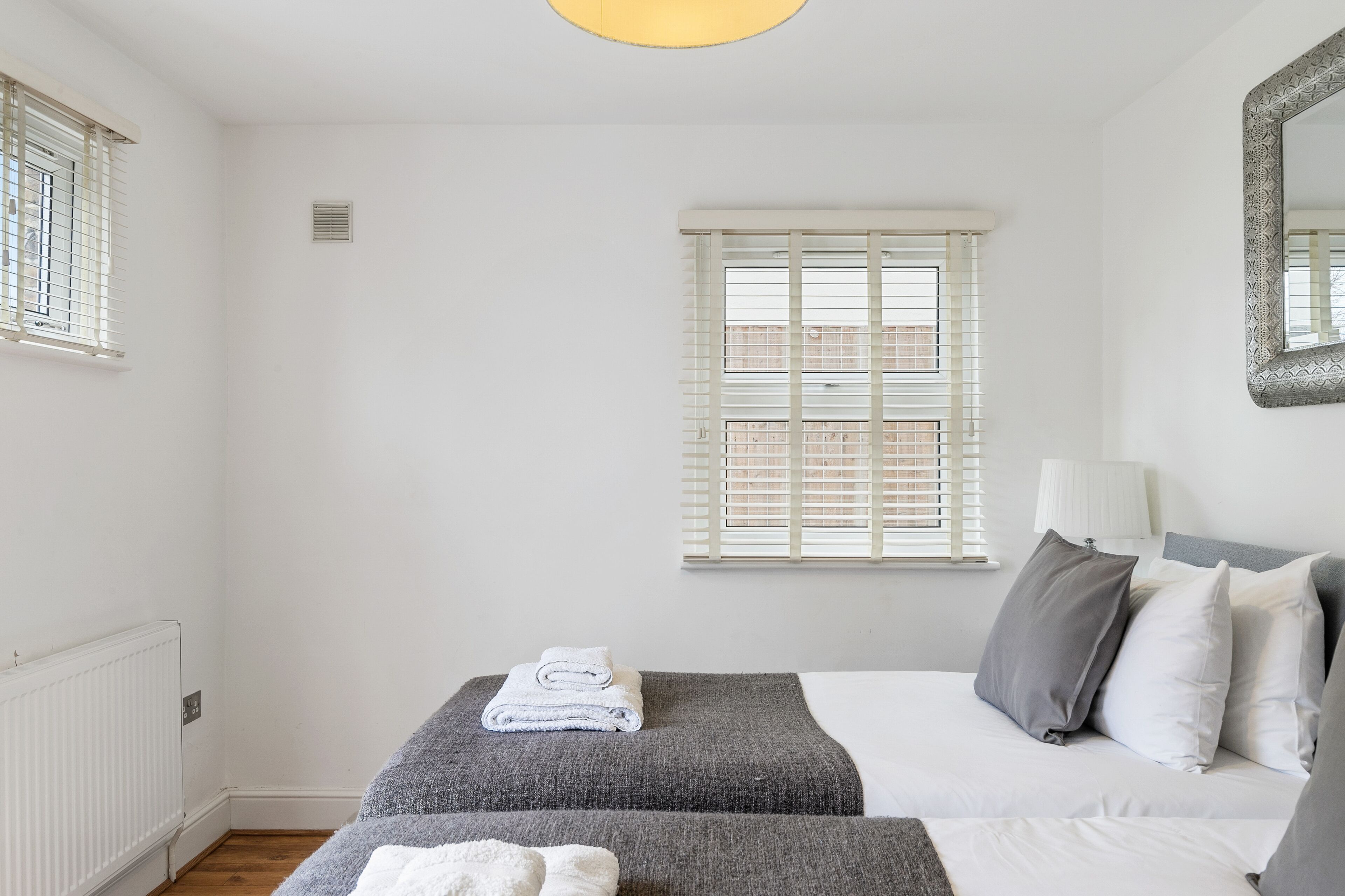 Une chambre avec deux lits, des murs blancs, un radiateur, une fenêtre avec des stores et une lampe de chevet.