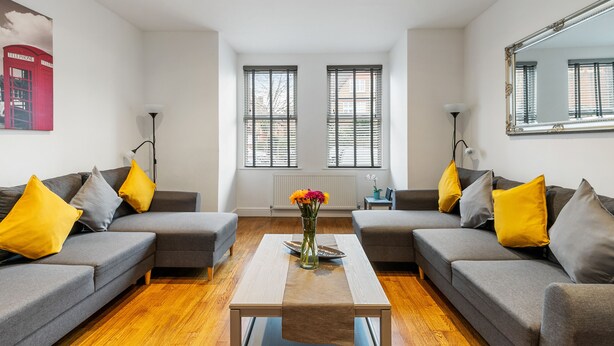 A modern living room with grey sofas, yellow and grey cushions, a central coffee table with a vase of flowers, and large windows with blinds. - St Marks Modern Apartments (Enfield)