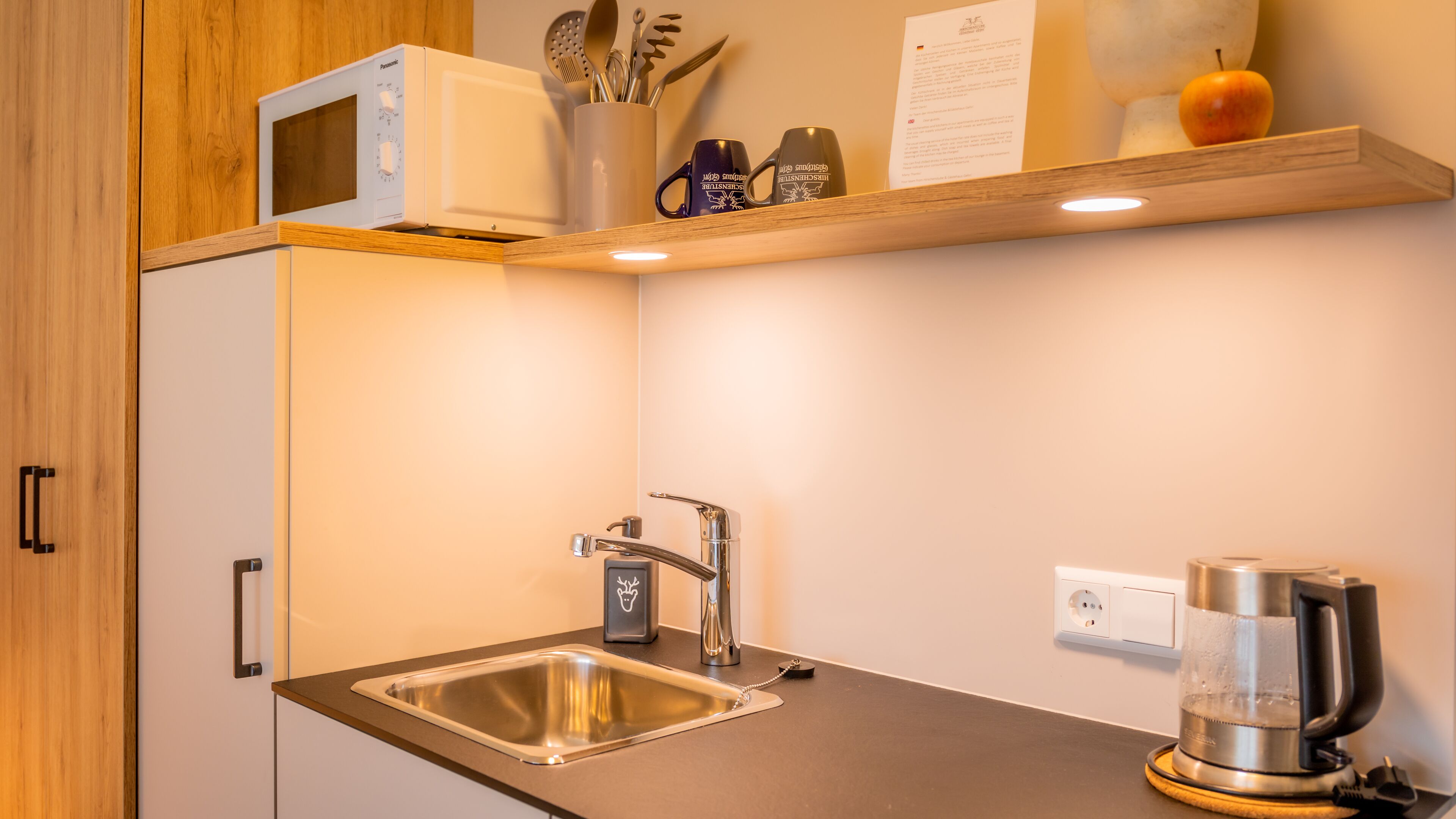 Una cocina con microondas integrado, pileta de acero inoxidable y tetera sobre la encimera.