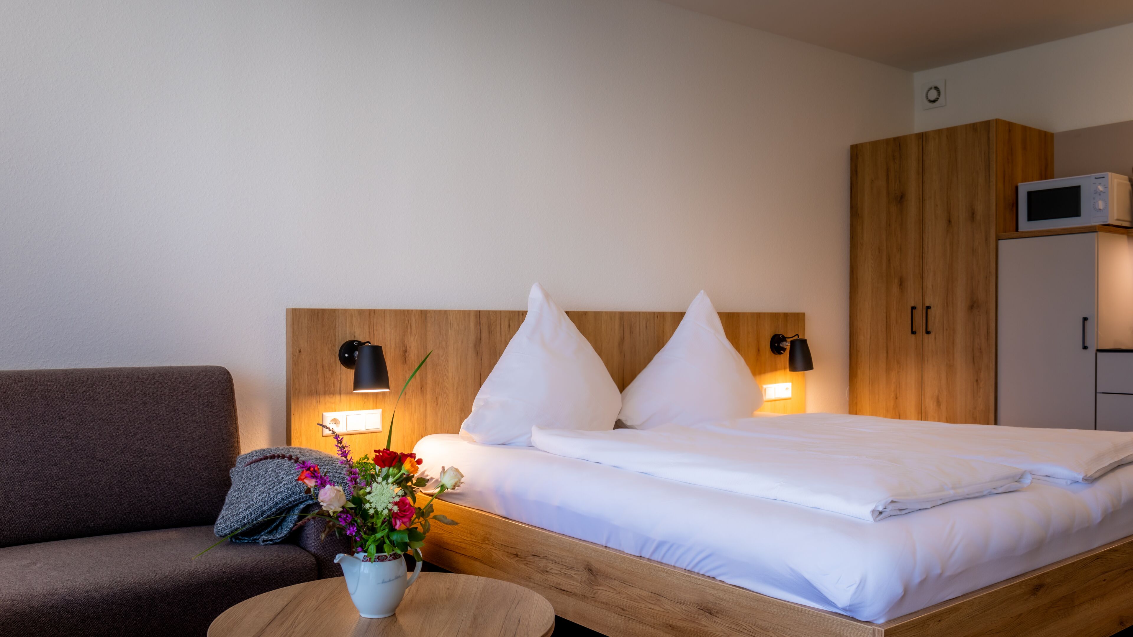 A hotel room with a bed, a wooden headboard, a wooden wardrobe, a microwave, a small table with a vase of flowers, and a brown sofa.