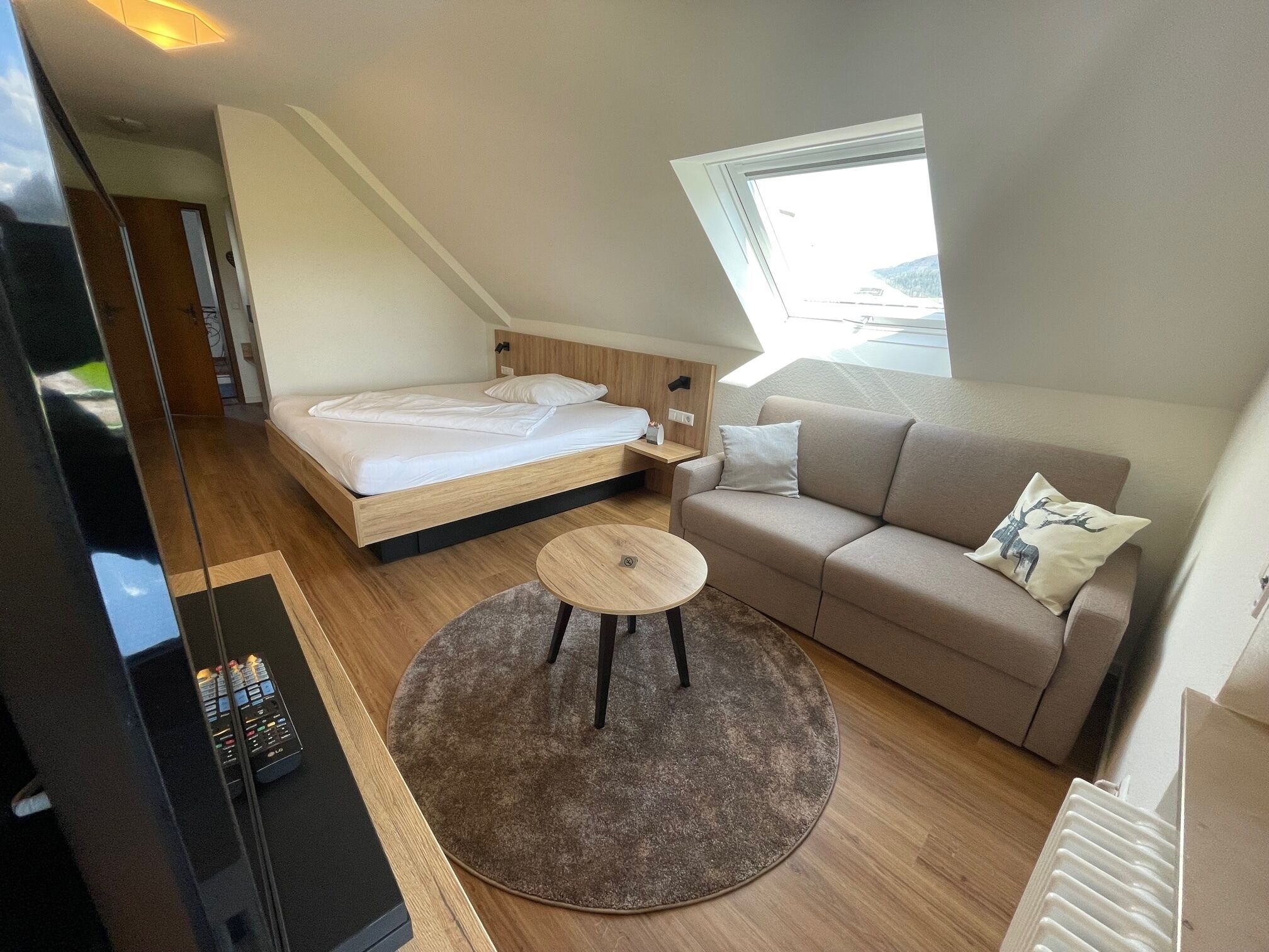 A modern, compact room with a bed, a sofa, a small round table, and a window providing a view of the outdoors.