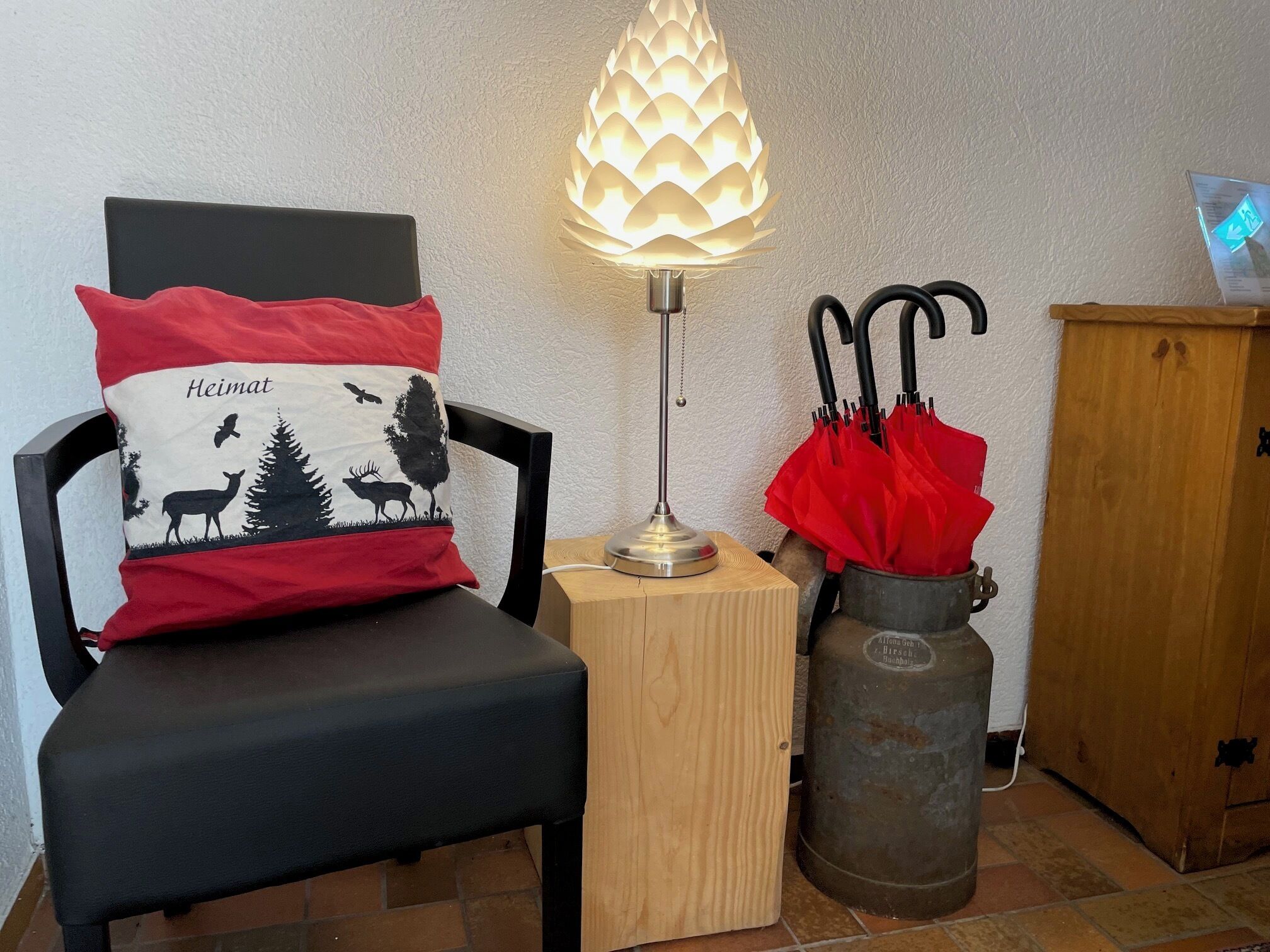 A chair with a pillow featuring a deer and trees, a table lamp, a metal bucket with a red umbrella, and a wooden cabinet.