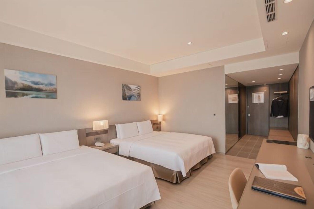 A hotel room with two beds, a desk, and a chair. There are wall-mounted lamps and artwork.