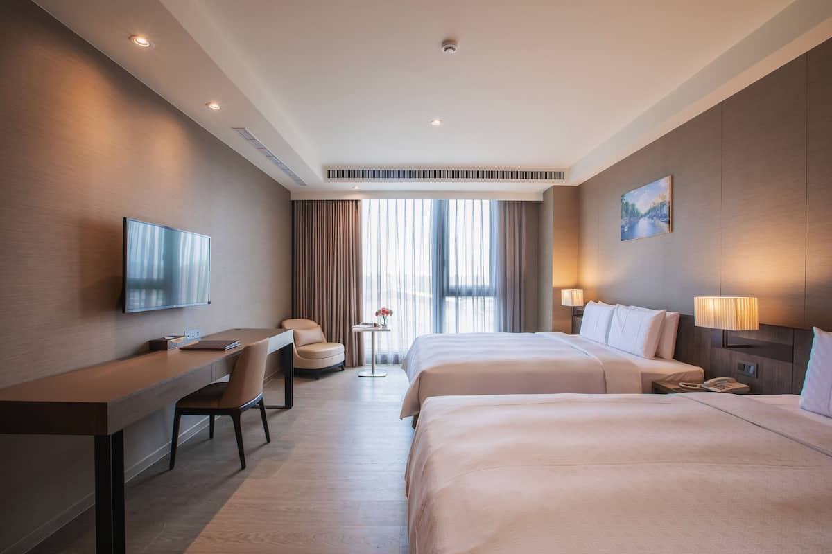 Ein Hotelzimmer mit zwei Betten, einem Schreibtisch, einem Stuhl, einem Fernseher, einem kleinen Tisch mit einer Vase und einem Bild an der Wand.