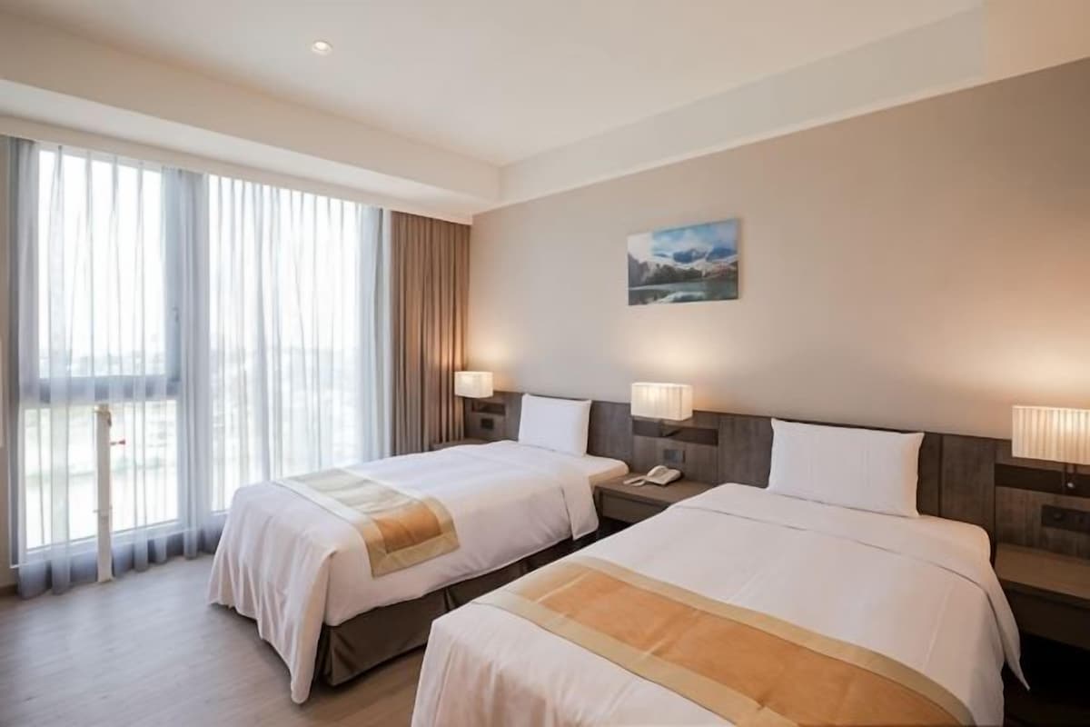 Quarto de hotel com duas camas, uma janela grande com cortinas, um quadro na parede e uma mesa de cabeceira com abajur.
