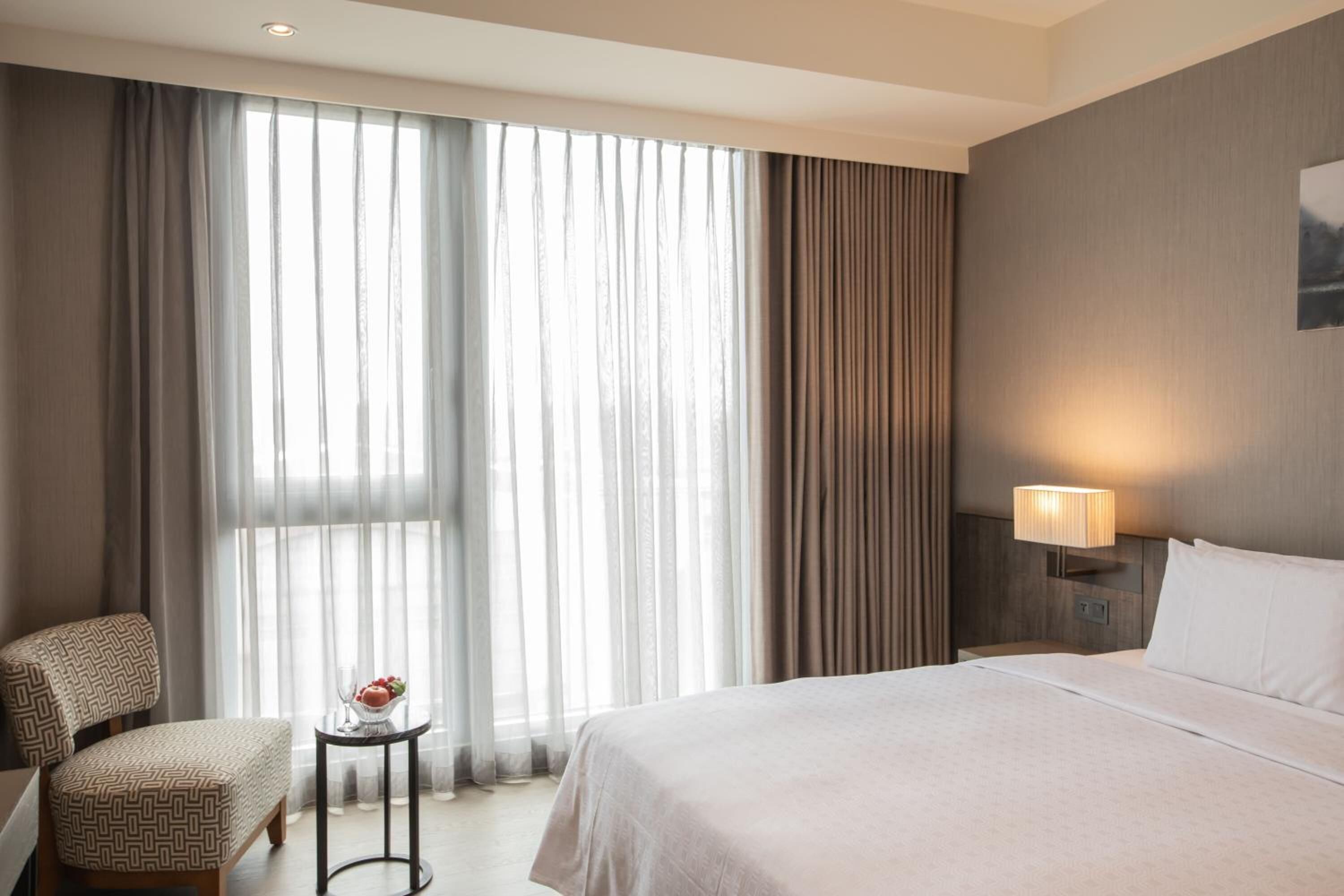 A hotel room with a large bed, a chair, a small table with a vase, and a lamp.