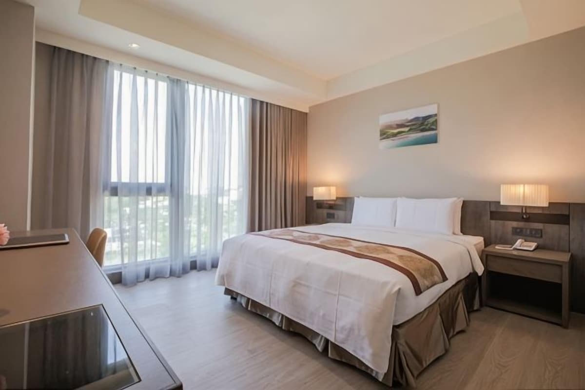 Quarto de hotel com uma cama grande, uma mesa de trabalho e vista para o exterior pela janela.