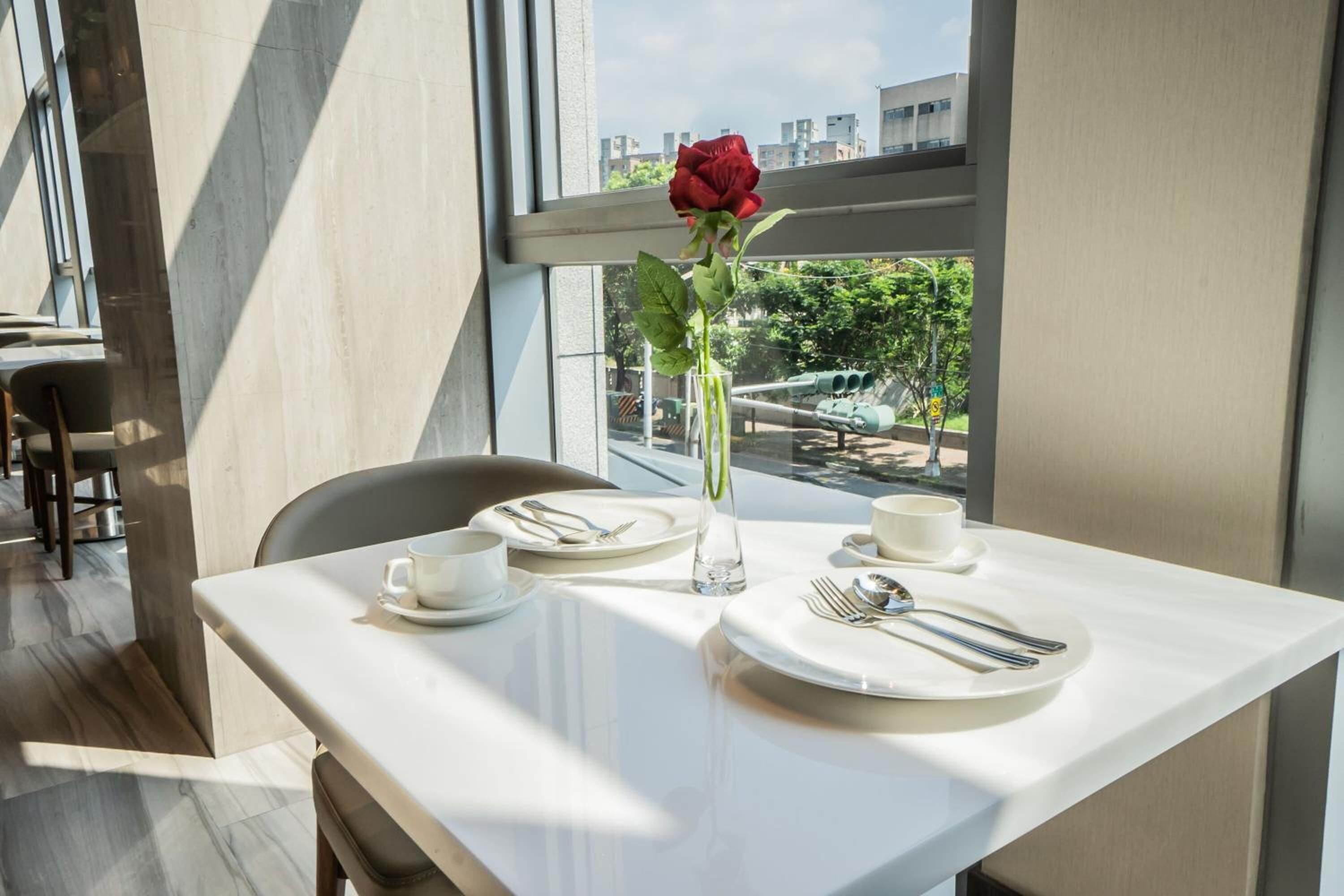 Une table à manger est dressée avec des assiettes, des couverts et un vase contenant une rose, le tout placé près d’une fenêtre offrant une vue sur des bâtiments et de la verdure.