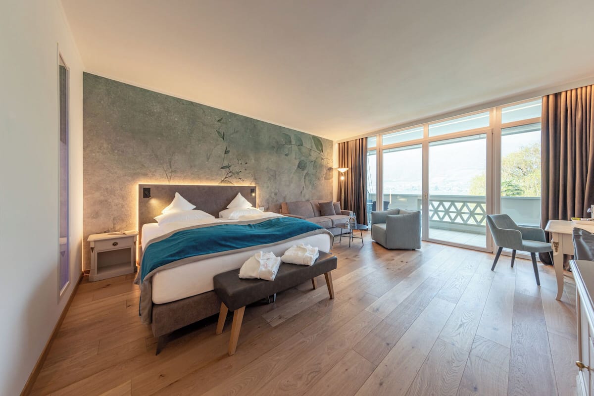 A modern hotel room with a large bed, a bench, a desk, and a balcony with a view.