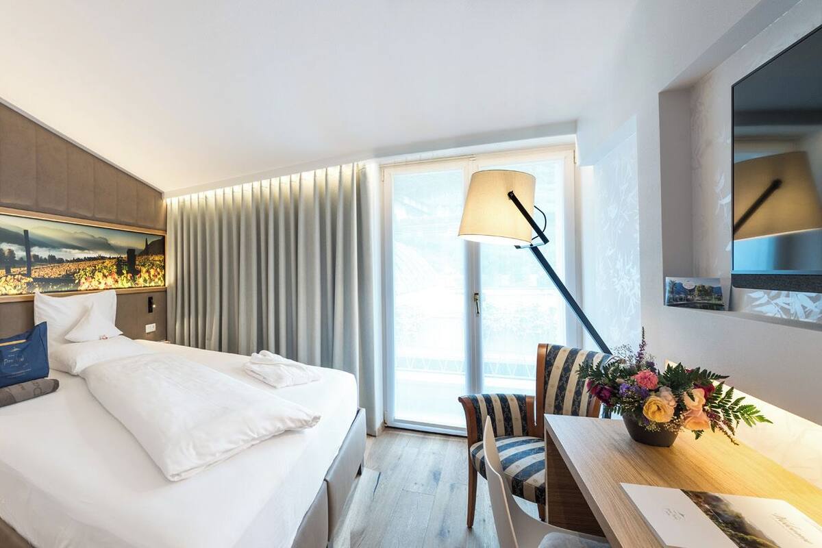 A hotel room with a bed, a desk with a lamp, a chair, a window with curtains, and a painting on the wall.