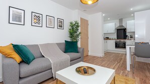 Een moderne woonkamer met een grijze bank, een witte salontafel en een eethoek met een grijze stoel. Achtergrond toont een keuken met witte kasten en een fornuis.