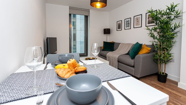 Een moderne woonkamer met een eethoek, met een grijze bank, een eettafel met een tafelkleed met dessin en een potplant.