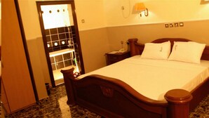 Ein Hotelzimmer mit einem hölzernen Bett, einem Nachttisch mit Lampe und einem durch eine Glastür einsehbaren Badezimmer.