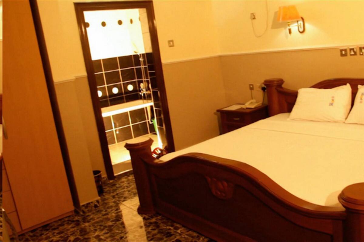 Hotellihuone, jossa on puinen sänky, yöpöytä lampun kanssa ja kylpyhuone, joka näkyy lasiovesta.