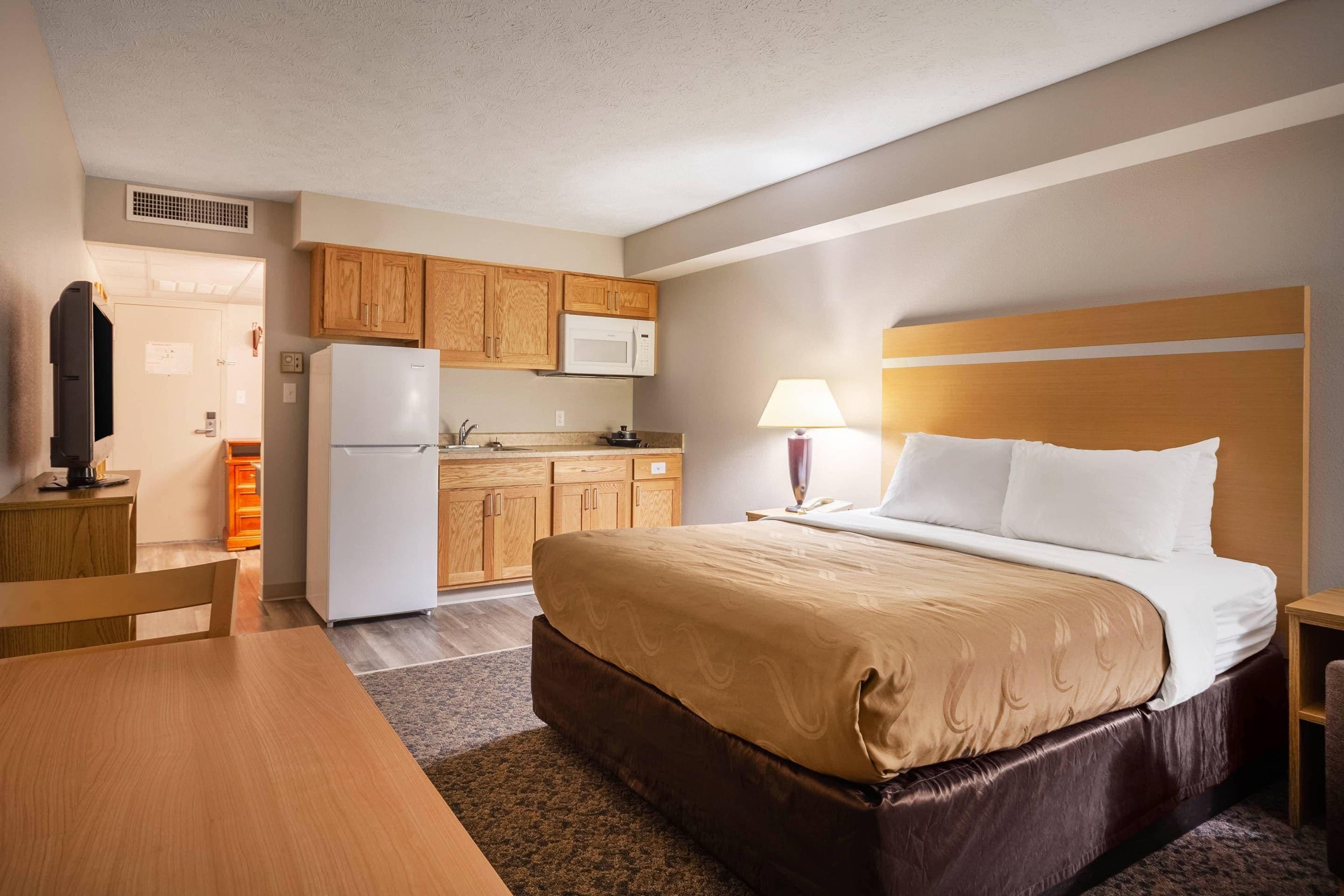A hotel room with a bed, a desk, a microwave, a refrigerator, wooden cabinets, and a small kitchenette.