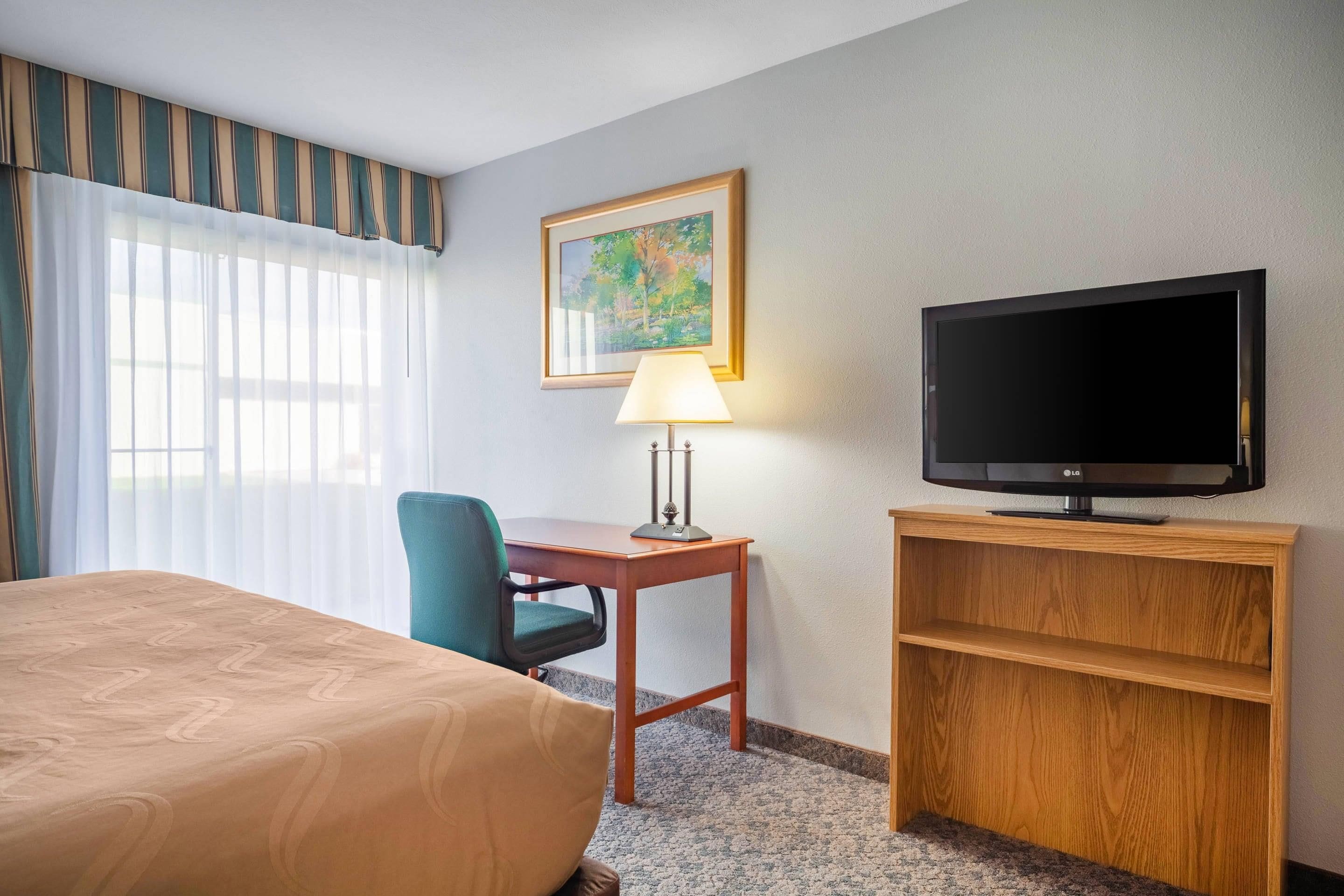 A hotel room with a bed, a desk with a lamp, a television, and a painting on the wall.