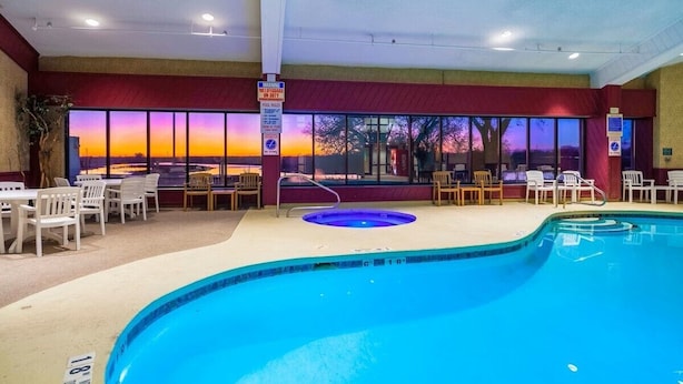 An indoor swimming pool with a sunset view through large windows, surrounded by white chairs and tables. - Suburban Studios I-80 Grand Island (Grand Island)