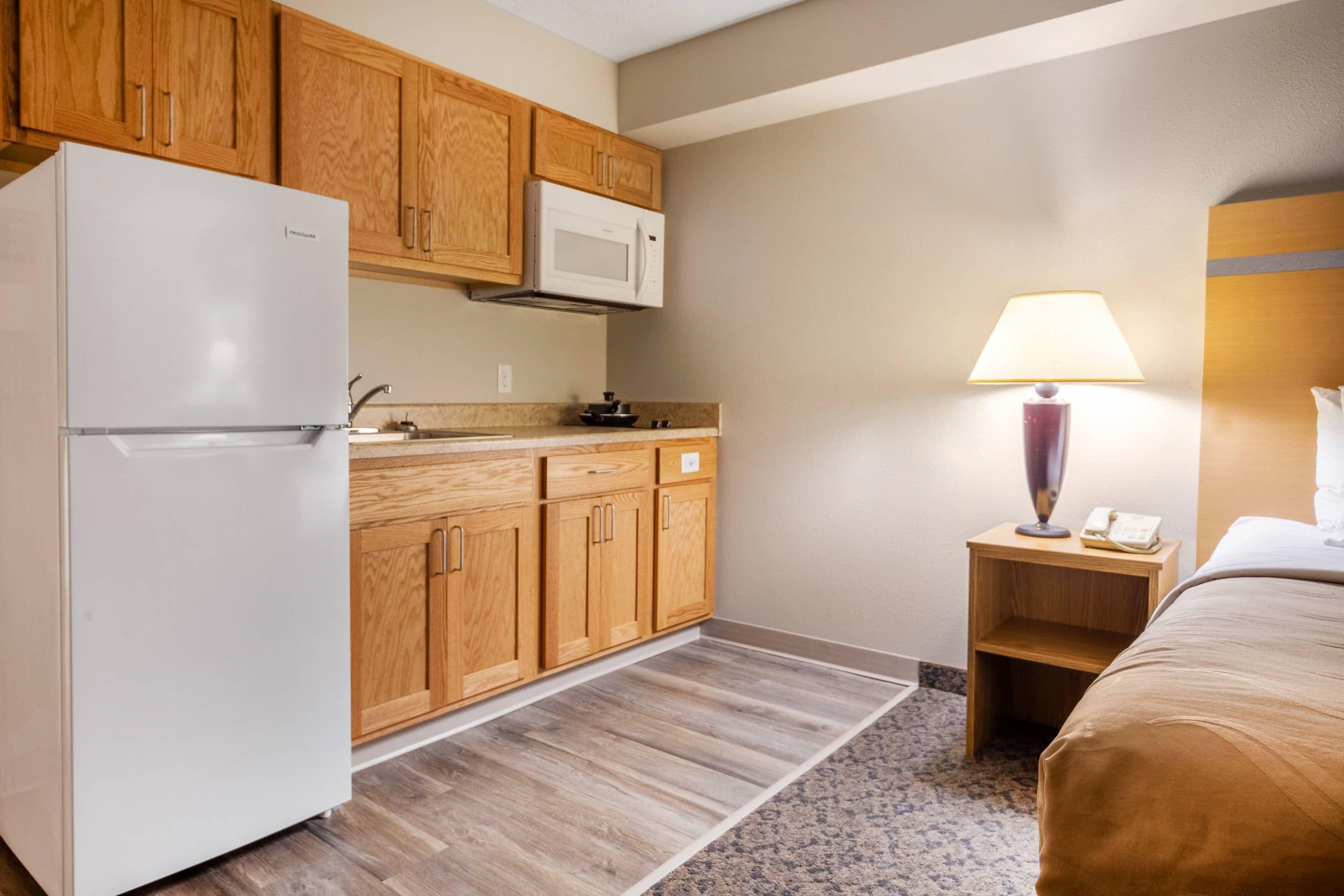 A compact hotel room with a kitchenette, including a refrigerator, microwave, and wooden cabinets. There's a bedside table with a lamp and a phone, and a bed with a headboard.