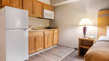 A compact hotel room with a kitchenette, including a refrigerator, microwave, and wooden cabinets. There's a bedside table with a lamp and a phone, and a bed with a headboard.