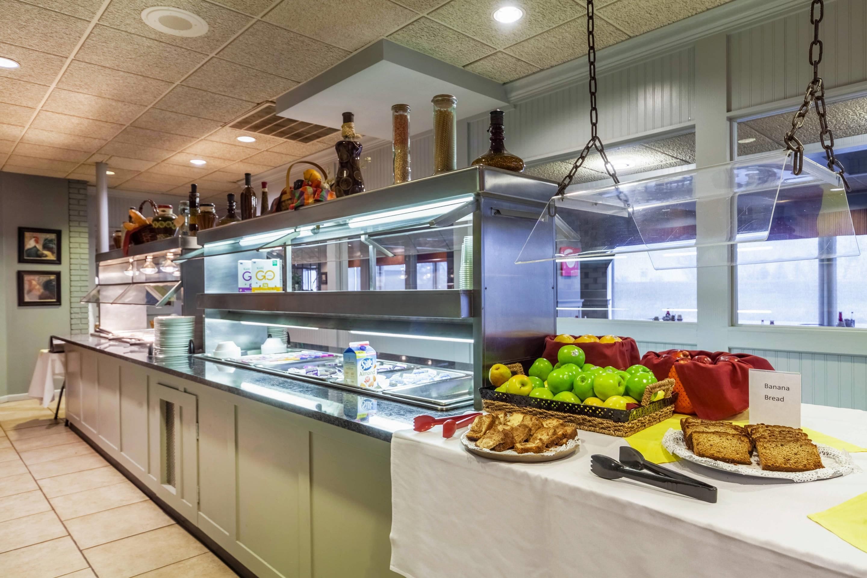 A buffet area with a glass display case containing various food items, a selection of fruits, and a sign indicating 'Banana Bread'.