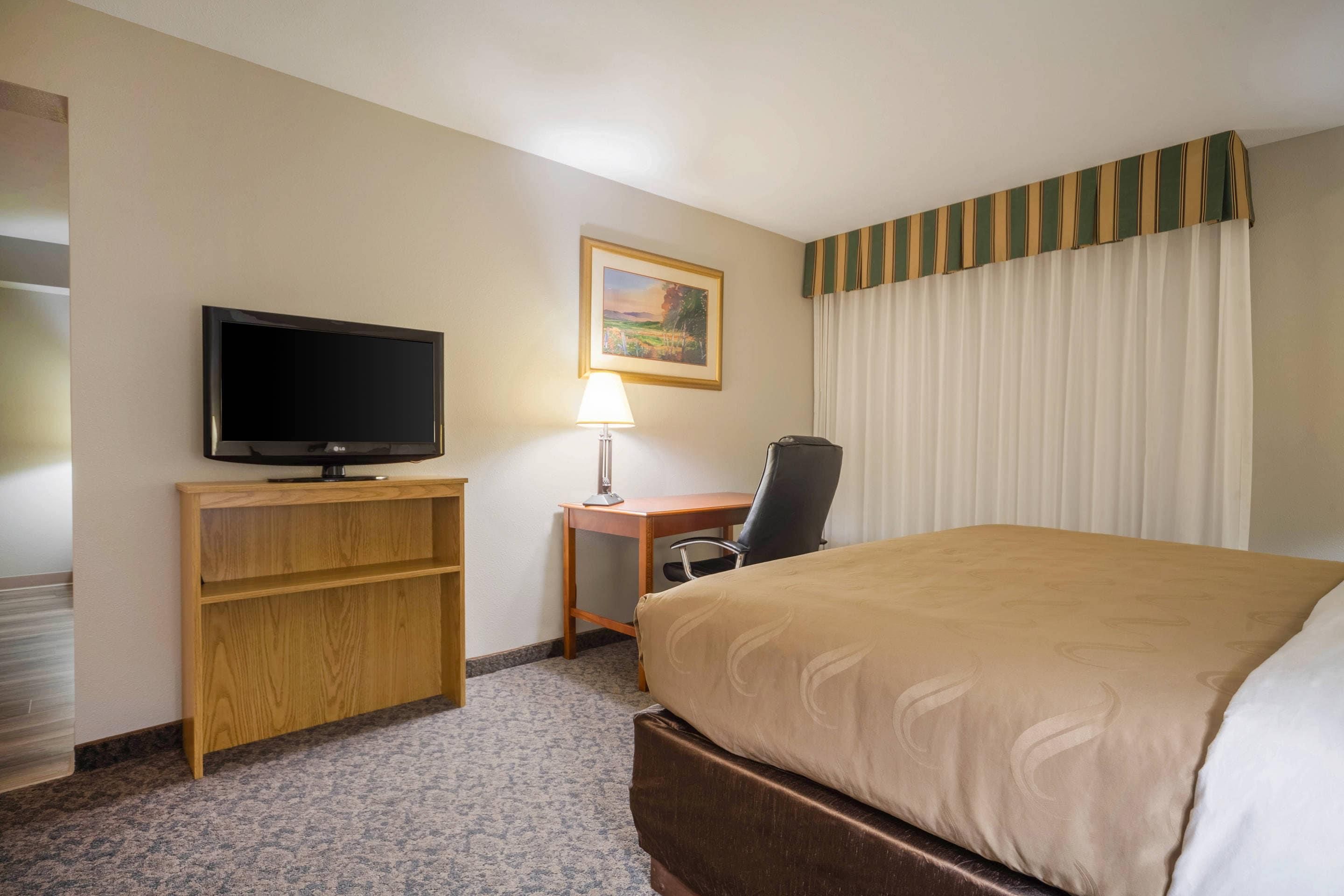 A hotel room with a bed, a desk with a chair, a television, a lamp, and a painting on the wall.