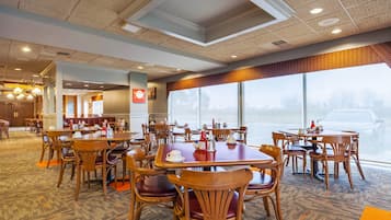 A hotel dining area with multiple tables and chairs, large windows, and a carpeted floor.