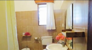 Et badeværelse med toilet, vask og spejl. Der er et vindue med gardin samt en hylde med blomster.