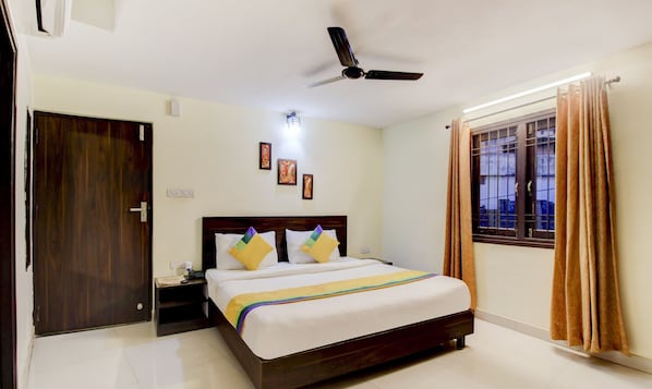 A hotel room with a bed, a ceiling fan, a nightstand, a window with curtains, and a door. - Itsy Hotels Royal Monarch (Gorakhpur)