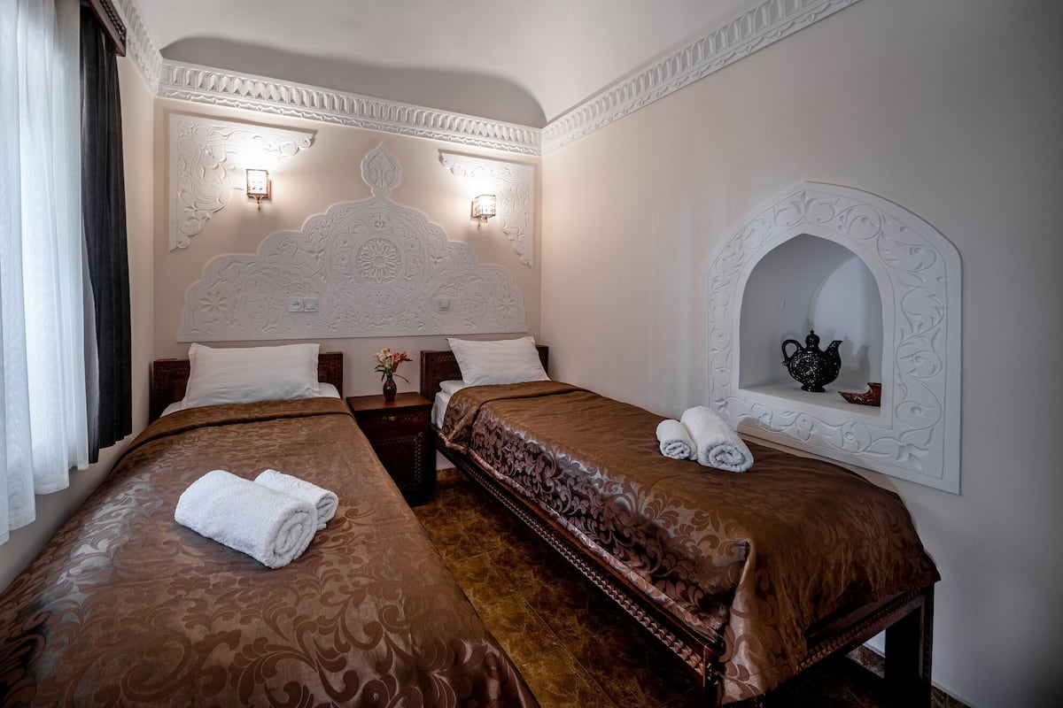 Une chambre d’hôtel avec deux lits, une tête de lit décorative, des appliques murales, un miroir avec un cadre orné et une petite table sur laquelle est posé un vase.
