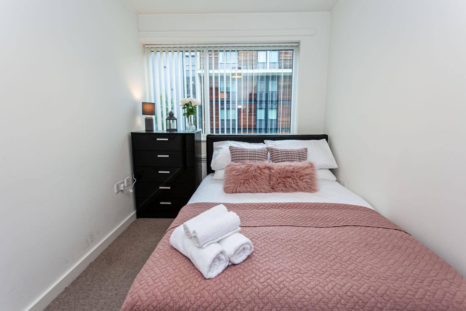 Un dormitorio con cama, cómoda, ventana con persianas y vista a edificios.