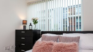 A bedroom with a bed, a dresser, a lamp, and a window with blinds. - Stunning Manchester Apartment (Salford)