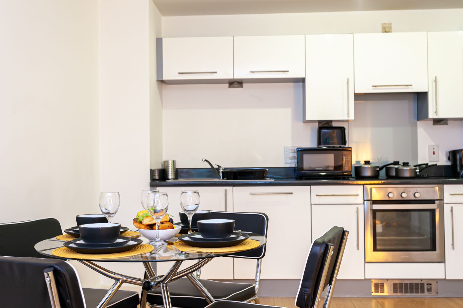 Una cocina moderna con armarios blancos, horno de acero inoxidable y una zona de comedor con mesa de vidrio y sillas negras.
