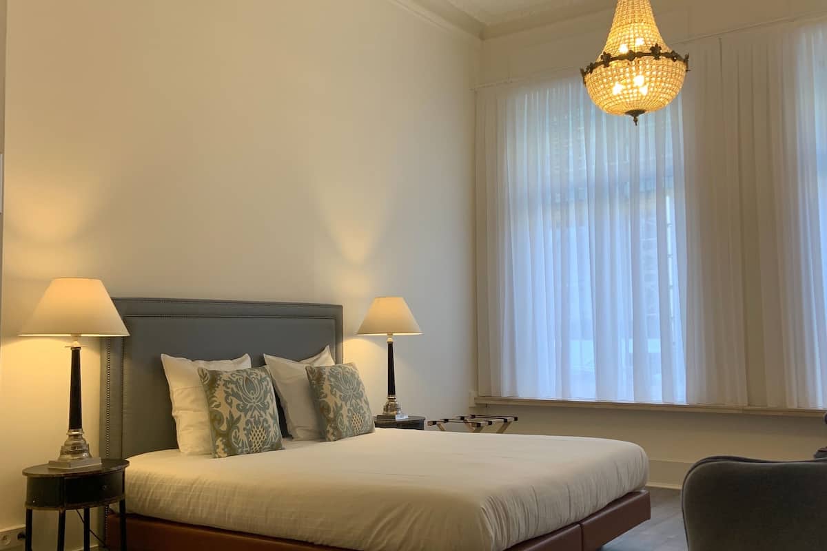 Une chambre spacieuse avec un grand lit, deux lampes de chevet, un lustre, une fenêtre avec des rideaux légers et un plancher en bois.