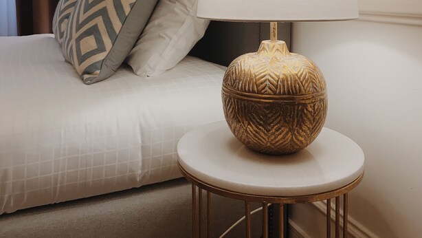 Un lit bien fait, avec un oreiller à motifs, une lampe de chevet posée sur une petite table ronde et une tête de lit.