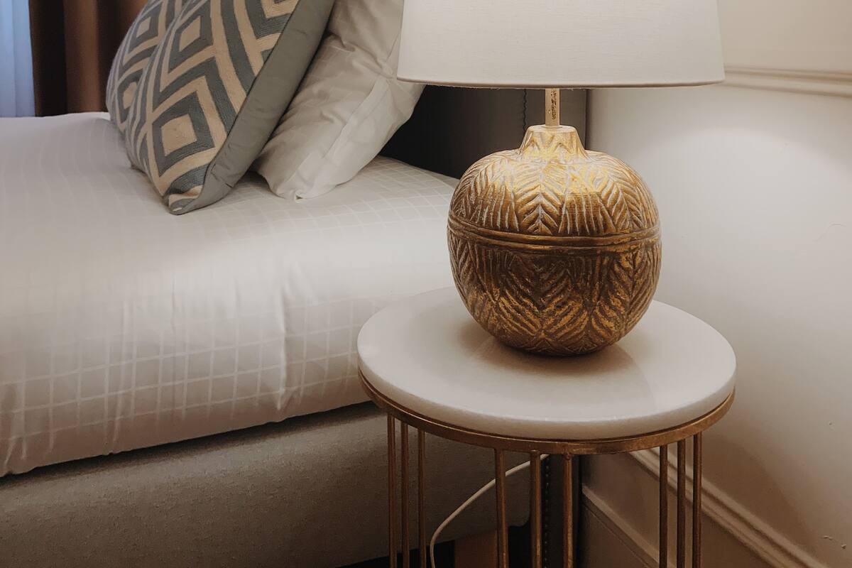 Un lit bien fait, avec un oreiller à motifs, une lampe de chevet posée sur une petite table ronde et une tête de lit.